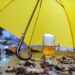 Volles Bierglas unter einem Regenschirm, der auf nassem Boden mit Herbstlaub steht.
