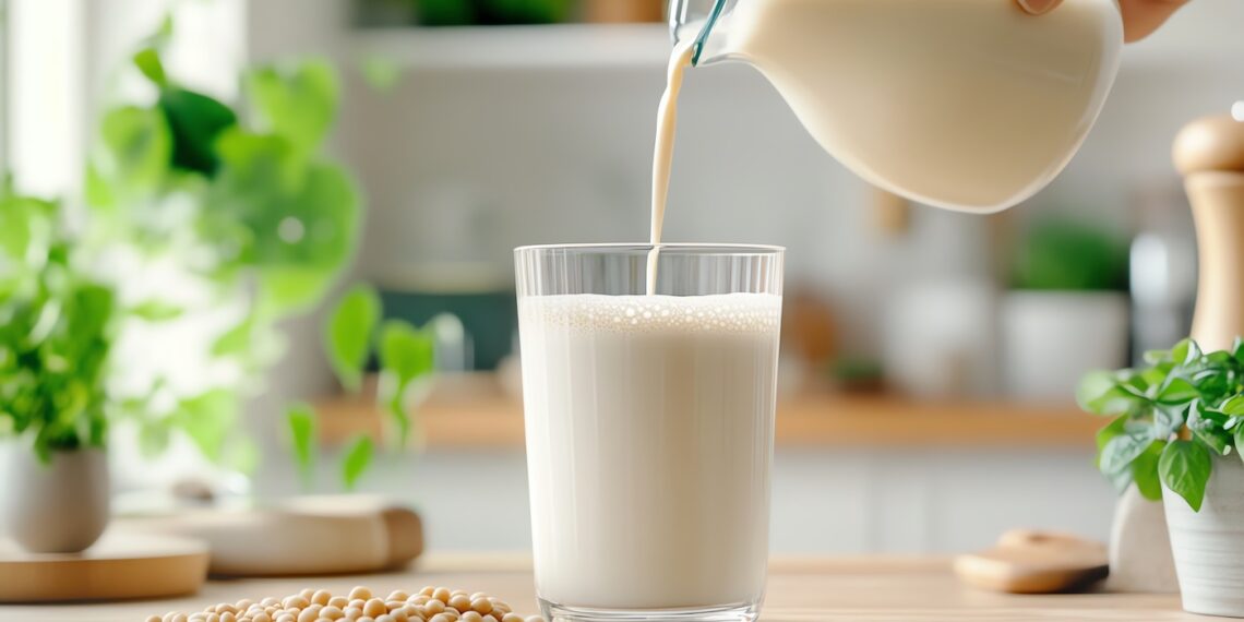 Eine Hand füllt Pflanzenmilch aus einem Krug in ein Glas, auf dem Tisch Pflanzen und Sojabohnen