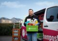 Ein lächelnder Flaschenpost-Fahrer steht vor seinem Lieferwagen und hält eine grüne Kiste mit Lebensmitteln. Neben ihm stehen Getränkekisten auf einem Rollwagen, im Hintergrund Wohnhäuser.