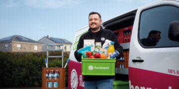 Ein lächelnder Flaschenpost-Fahrer steht vor seinem Lieferwagen und hält eine grüne Kiste mit Lebensmitteln. Neben ihm stehen Getränkekisten auf einem Rollwagen, im Hintergrund Wohnhäuser.