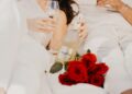 Zwei Menschen in heller Kleidung genießen gemeinsam ein Glas Freixenet-Sekt. Eine Flasche Freixenet liegt locker in der Hand, daneben ein Strauß roter Rosen.