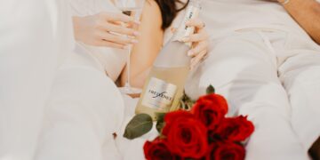 Zwei Menschen in heller Kleidung genießen gemeinsam ein Glas Freixenet-Sekt. Eine Flasche Freixenet liegt locker in der Hand, daneben ein Strauß roter Rosen.