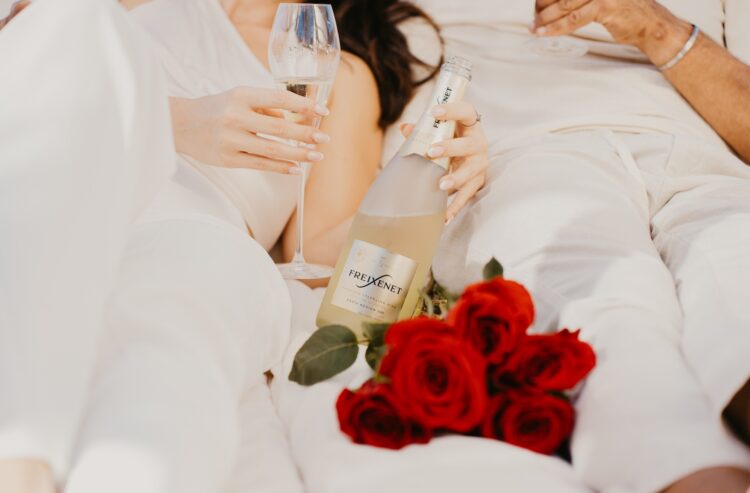 Zwei Menschen in heller Kleidung genießen gemeinsam ein Glas Freixenet-Sekt. Eine Flasche Freixenet liegt locker in der Hand, daneben ein Strauß roter Rosen.