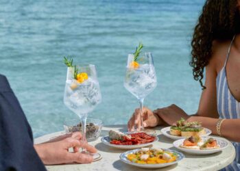 Ein Mann und eine Frau an einem runden Tisch mit zwei Gläsern Gin Tonic und kleinen mediterranen Speisen, im Hintergrund das Meer