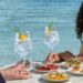 Ein Mann und eine Frau an einem runden Tisch mit zwei Gläsern Gin Tonic und kleinen mediterranen Speisen, im Hintergrund das Meer