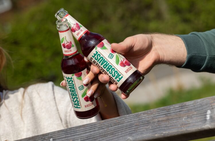 Foto von zwei Händen, die je eine Flasche Krombacher's Fassbrause Kirsche halten und damit anstoßen